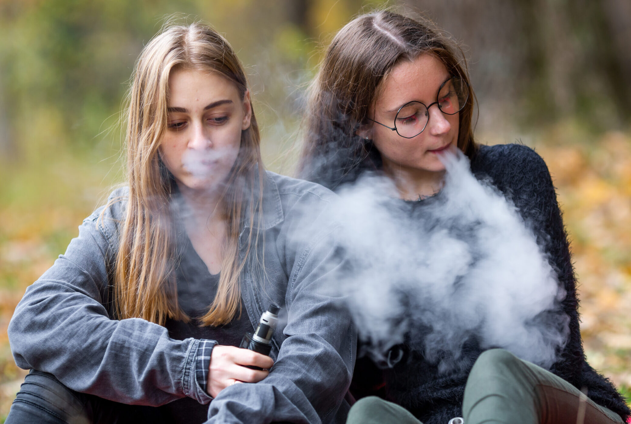 Vape lgbt teenagers. Bisexual lesbian young caucasian teenage girls in casual clothes vape electronic cigarette on the street in the park in the autumn evening. Bad habit.