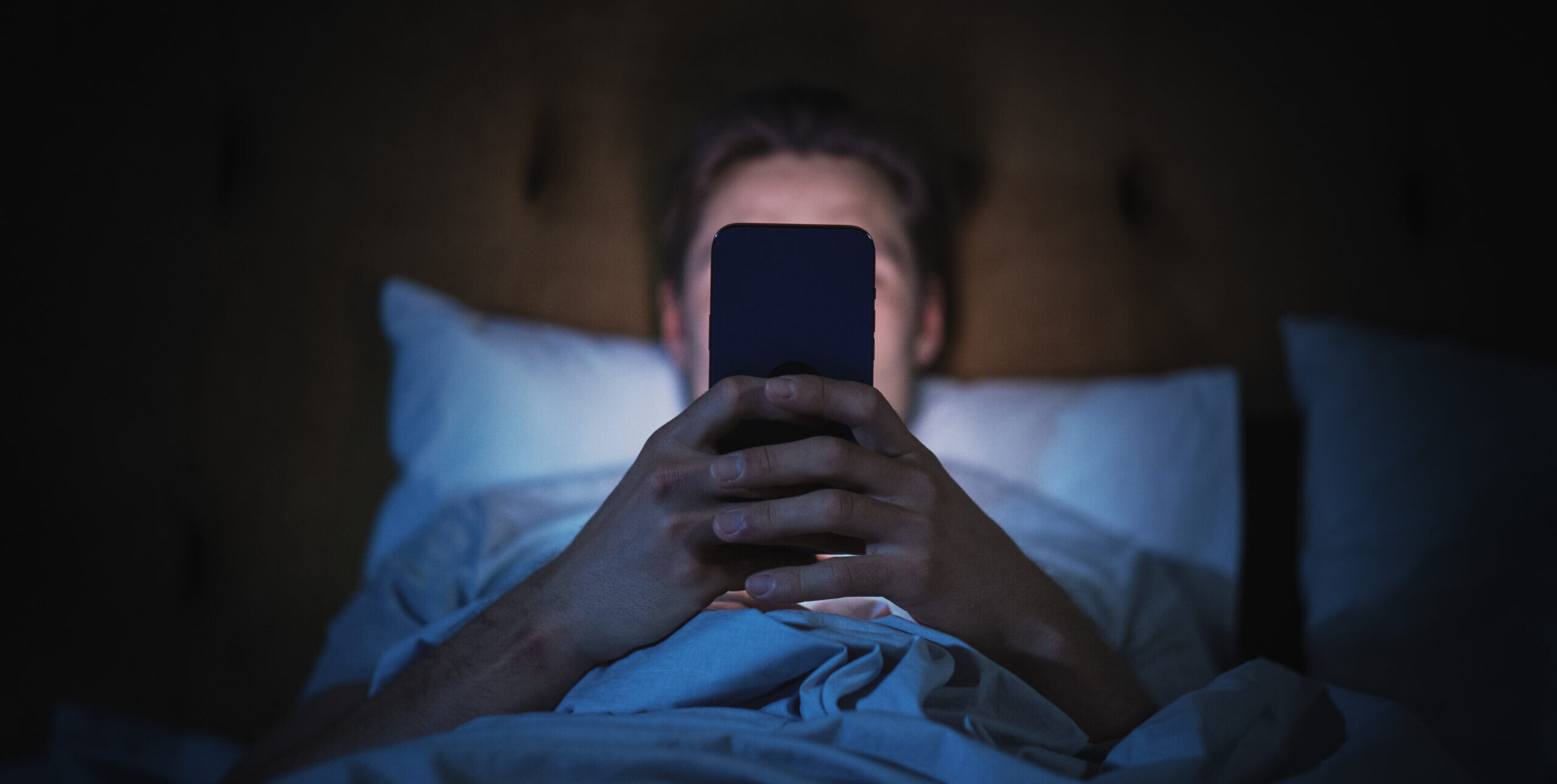 Anonymous Man Uses Smartphone in Bed at Home at Night. Handsome Guy Browsing Social Media, Reading News, Doing Online Shopping Late at Night. Focus on Hand Holding Mobile Phone Covering Face