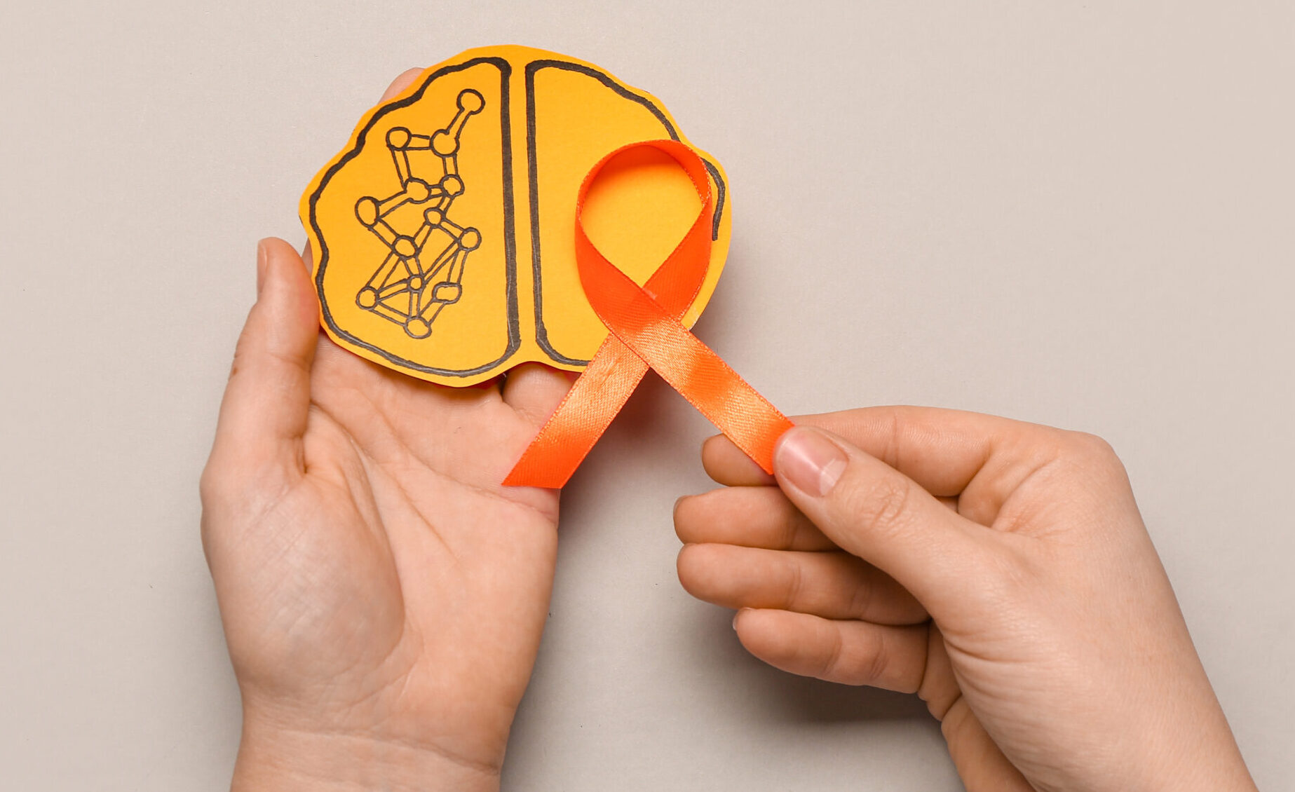 Female hands with paper brain and orange ribbon on grey background. Multiple Sclerosis Awareness Month